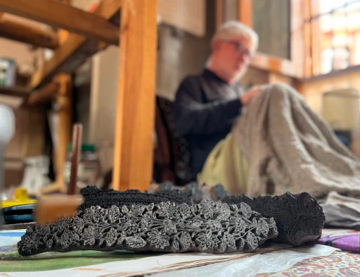 Hand-carved wooden printing blocks displayed in the foreground while an artisan works on handcrafted Kashmiri textiles in a traditional workshop.