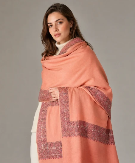 Model wearing a handcrafted Kashmiri embroidered shawl in peach with intricate paisley and floral embroidered borders and a soft woven finish.