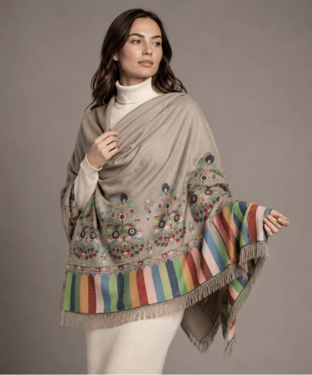Model wearing a handcrafted Kashmiri woven shawl in taupe featuring colorful floral motifs, multicolor striped borders, and soft fringed edges.